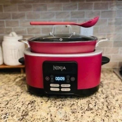 Fuchsia Ninja cooking appliance with a digital display, featuring a matching pan, glass lid, and ladle, set on a granite countertop with a tiled kitchen backsplash.