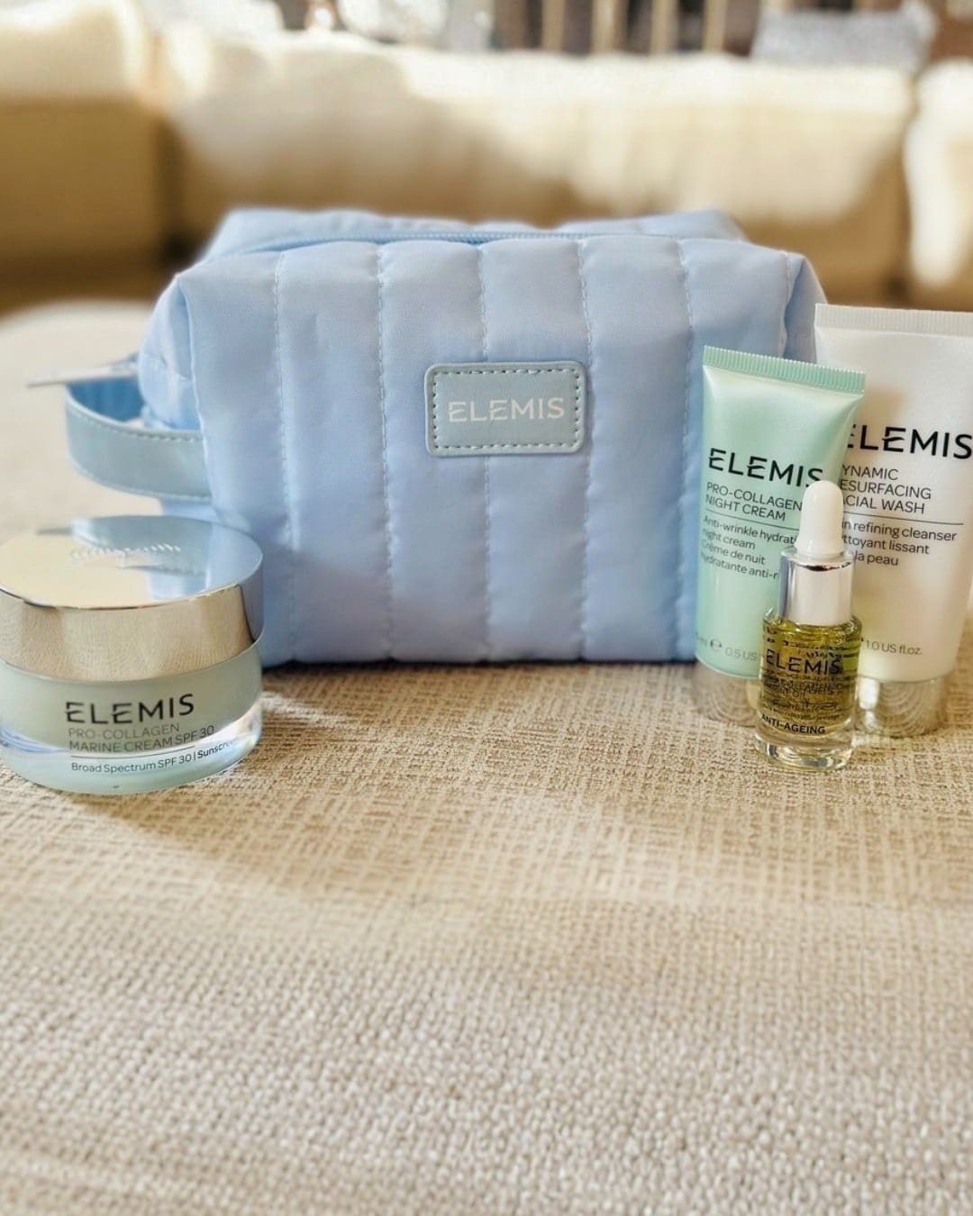 Elemis cream set featuring a light blue quilted toiletry bag with Pro-Collagen Marine Cream SPF 30, Pro-Collagen Night Cream, Dynamic Resurfacing Facial Wash, and anti-aging oil, arranged on a textured surface in a cozy indoor setting.