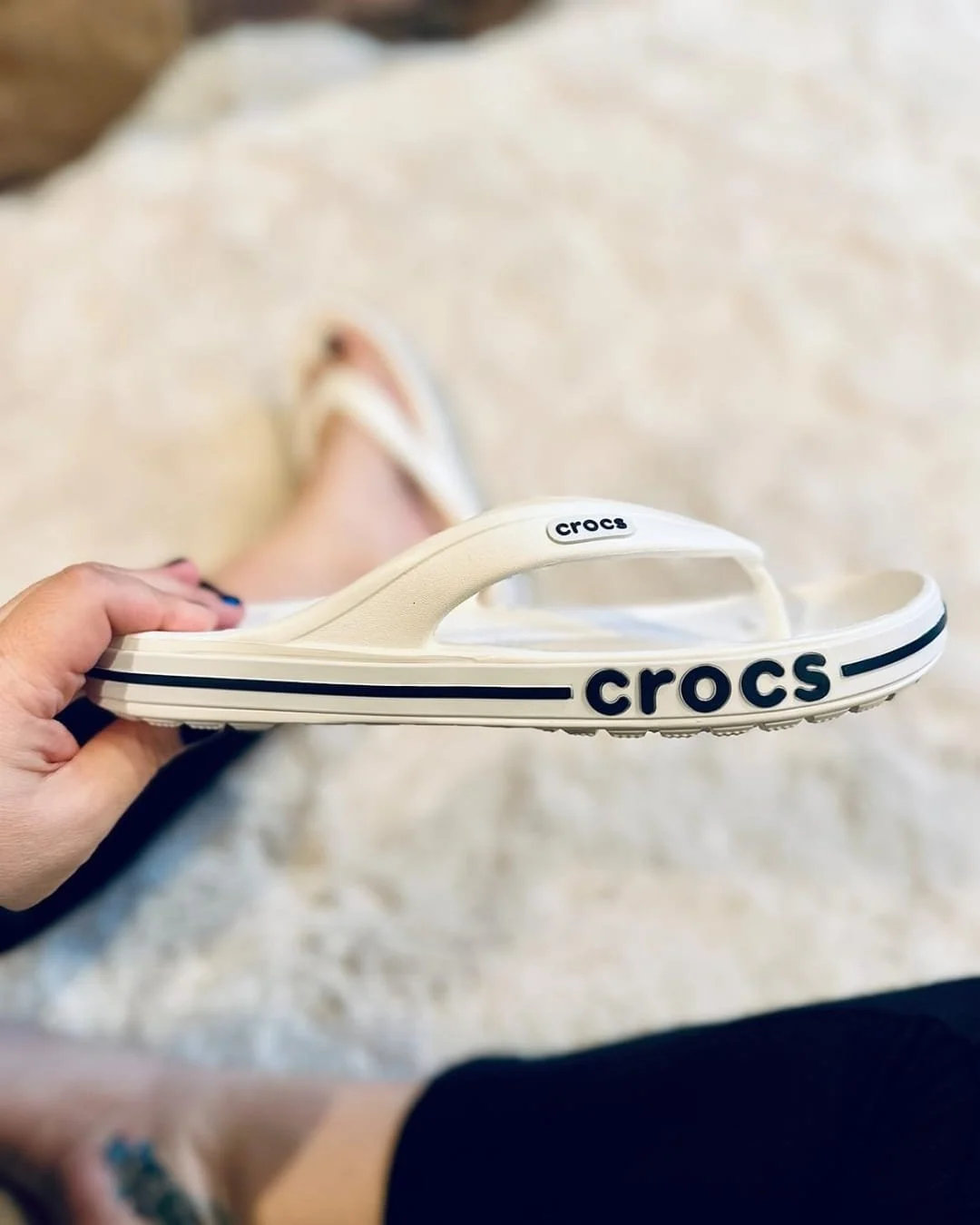 Close-up of a white Crocs Crocband Flip sandal with dark blue accents, held in a hand over a soft white textured surface, with the wearer's feet and a tattooed ankle visible in the background.