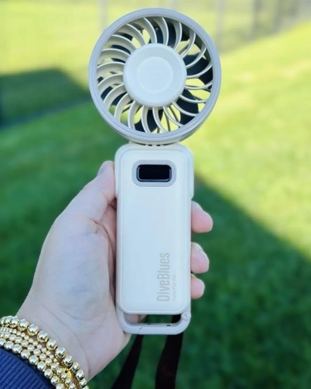 Hand holding a cream-colored DiveBlues Turbo Fan Pro handheld fan with a digital display and wrist strap, shown outdoors against a sunny grassy background.