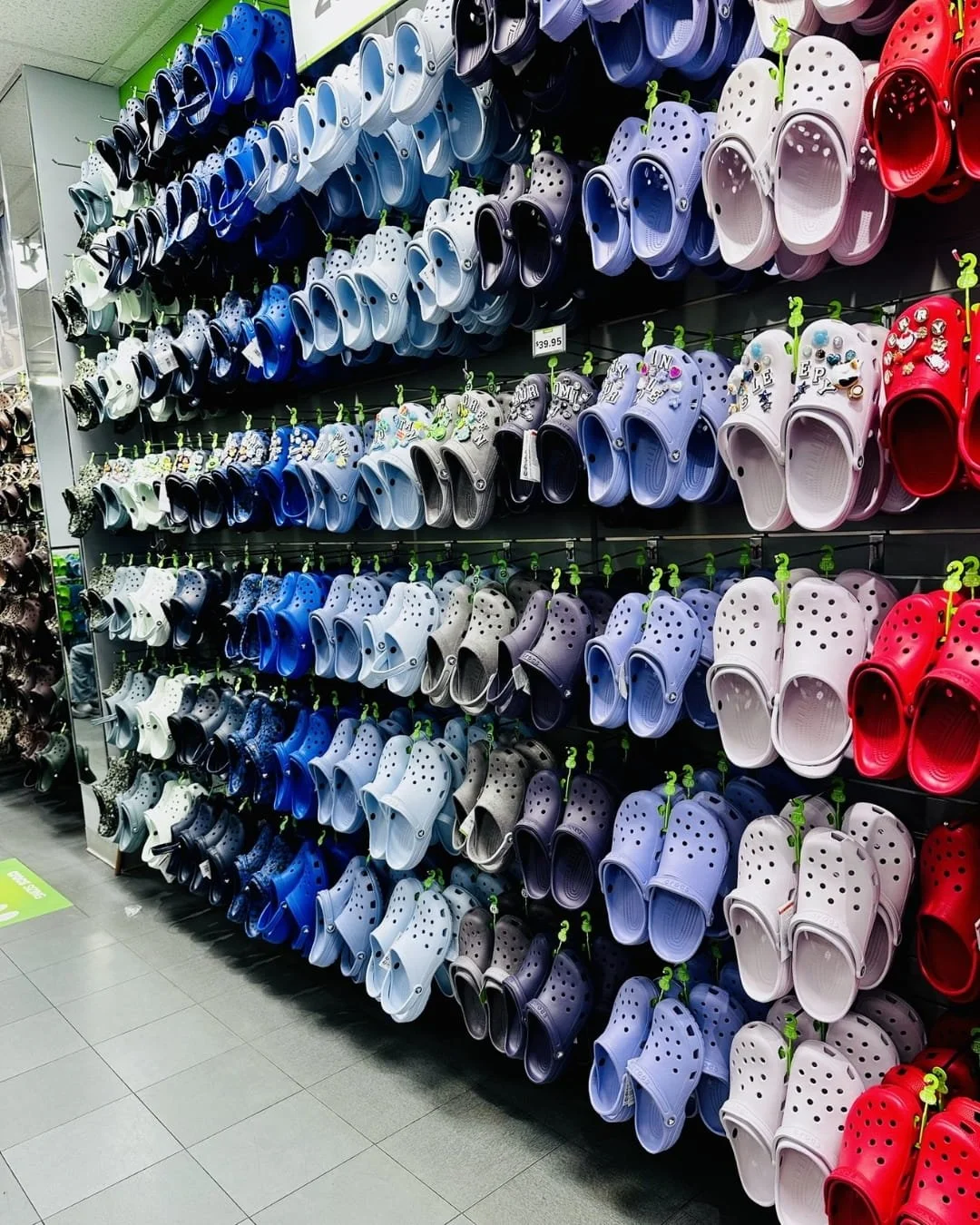 Color-coordinated retail display of Crocs clogs and sandals arranged on a slatwall, featuring a gradient of colors from dark blue to red, with Jibbitz charms on select pairs.