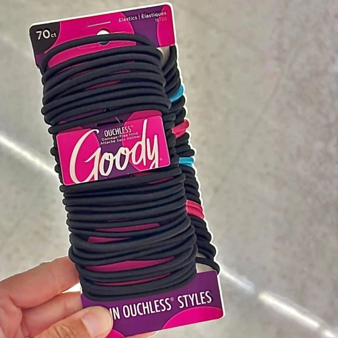 Goody Ouchless black hair elastics, 70-count pack with damage-free hold, displayed in vibrant fuchsia and purple packaging, held by a hand in a retail setting.