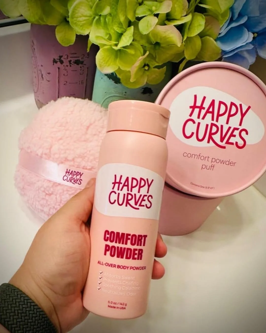 Happy Curves Comfort Powder and Powder Puff set in soft pink packaging, displayed on a white countertop with green hydrangeas and vintage glass jars in the background, emphasizing a feminine and self-care aesthetic.