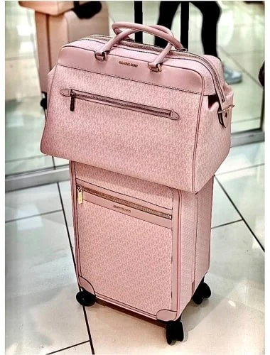 Light pink Michael Kors luggage set with a monogrammed handbag atop a matching rolling suitcase, featuring rose gold hardware, displayed on a tiled floor in a bright indoor setting.