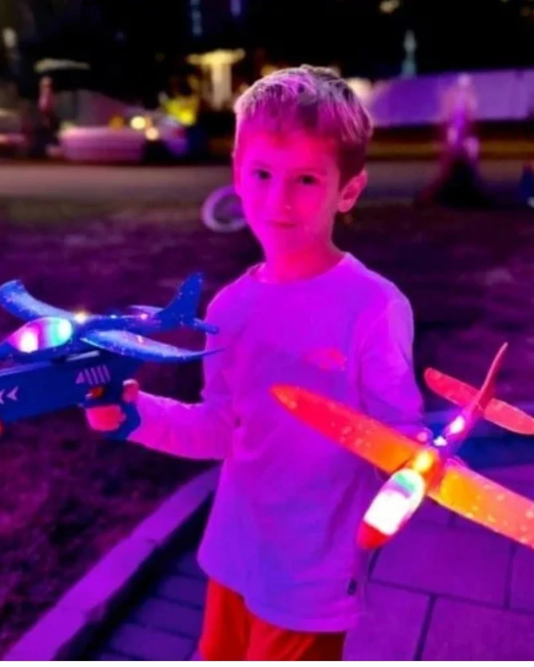 Young boy outdoors at night holding two light-up toy airplanes, one blue on a launcher and one orange, illuminated by vibrant purple lighting with a blurred dark background.