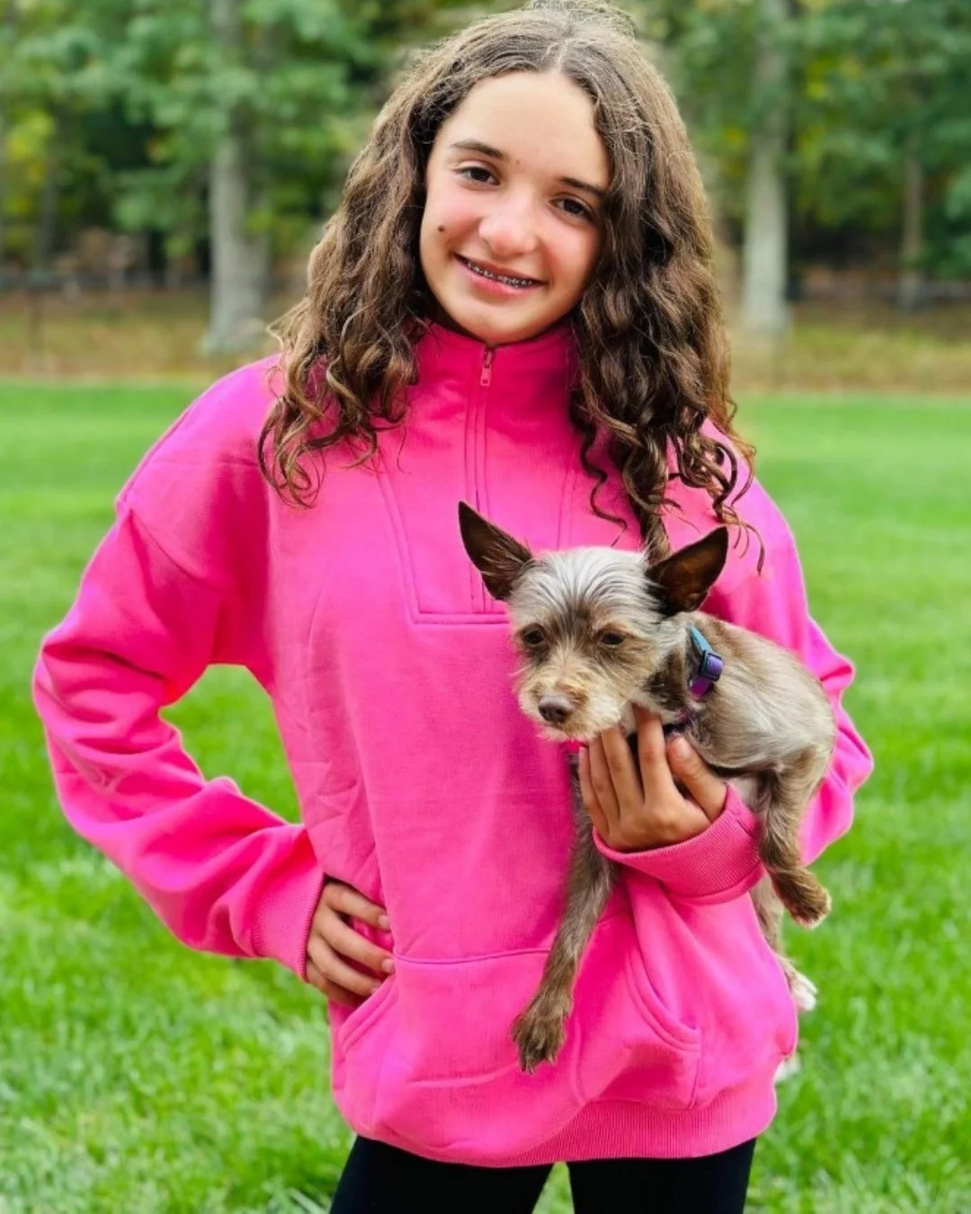 Smiling young girl with long curly brown hair wearing a vibrant hot pink half-zip sweatshirt, holding a small scruffy dog with grey and tan fur, standing outdoors on a green lawn with trees in the background.