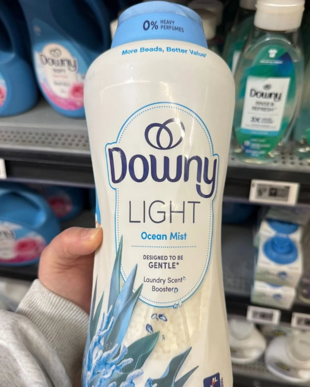Hand holding a white bottle of Downy Light Ocean Mist Laundry Scent Booster with a light blue cap, featuring gentle fragrance branding and visible scent beads, displayed in a retail laundry aisle.