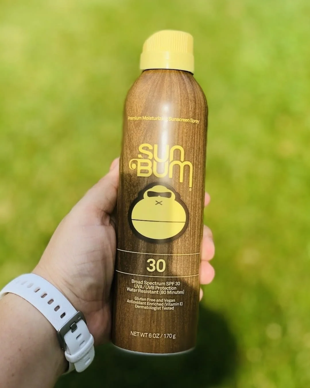 Hand holding a 6 oz Sun Bum Premium Moisturizing Sunscreen Spray with SPF 30, featuring a wood-grain design and yellow monkey logo, against a blurred green grass background.