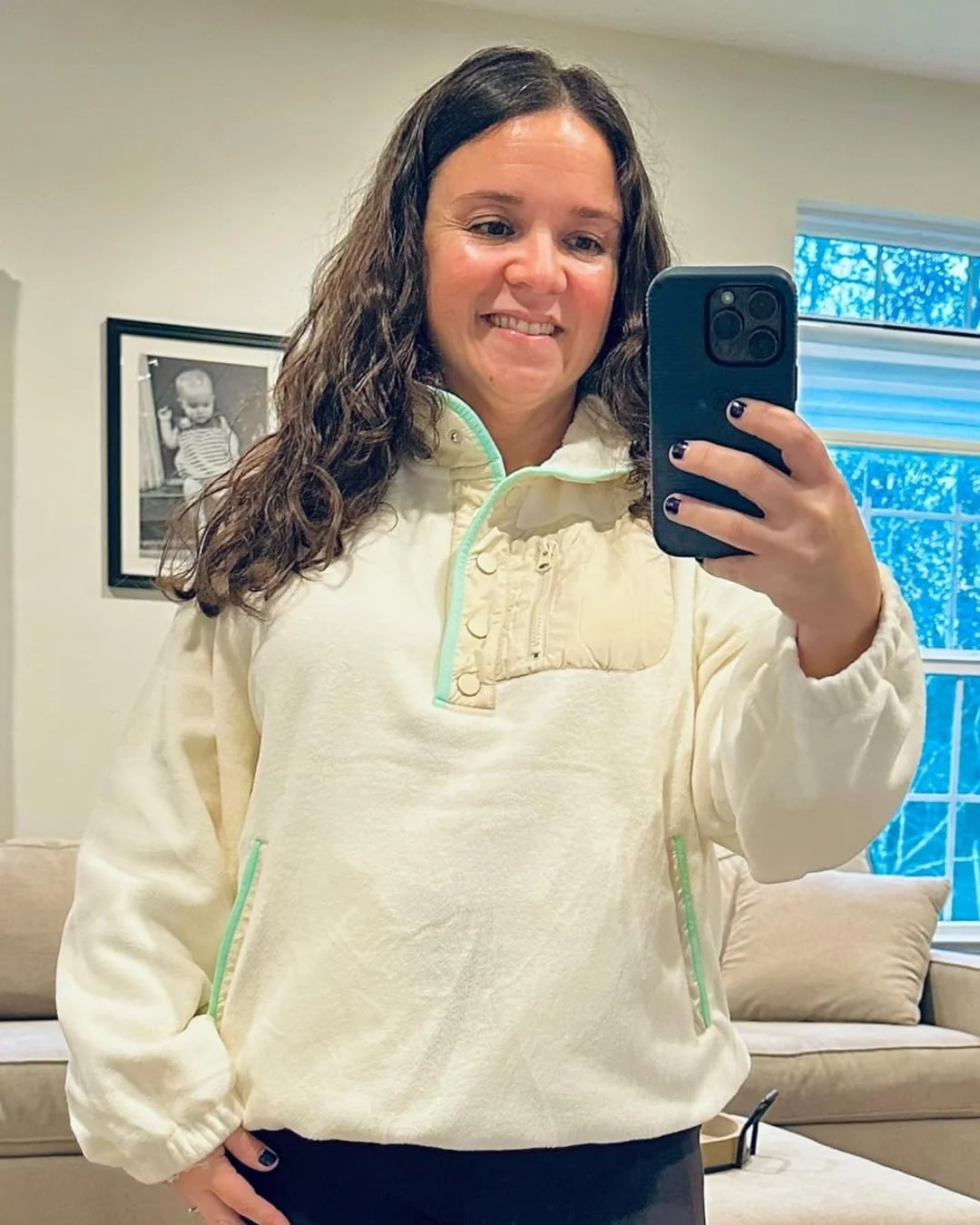 Nicole taking a mirror selfie wearing a cream-colored fleece pullover with mint green accents and tan quilted detailing, standing in a cozy room with a sofa, framed photo, and window in the background.