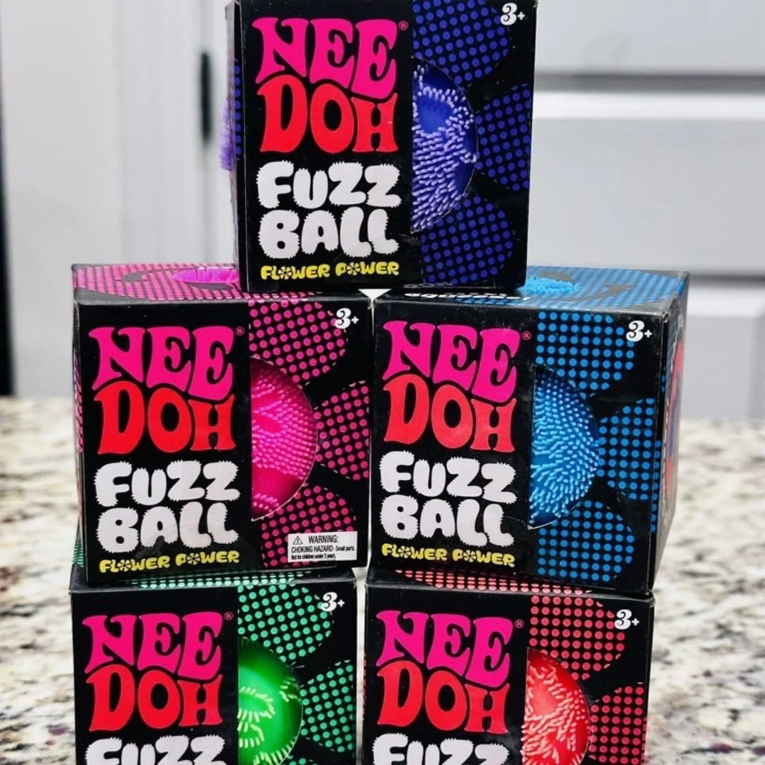 Stacked retail boxes of Nee Doh Fuzz Ball toys in vibrant colors—purple, pink, blue, green, and orange—featuring retro-style packaging with circular windows, displayed on a speckled countertop.