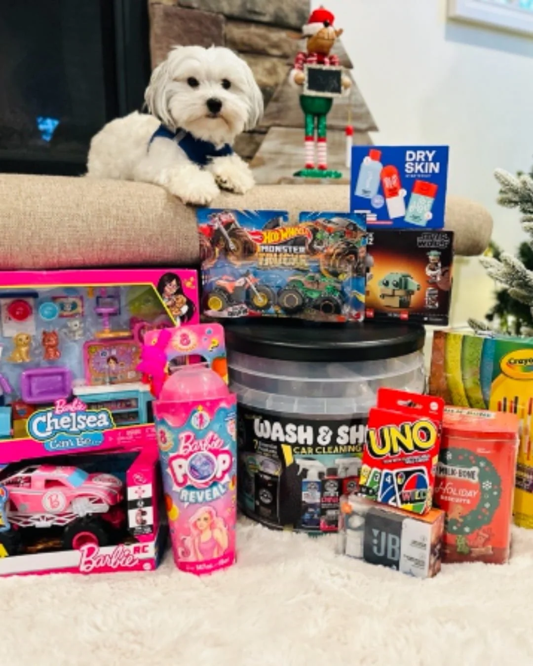 Holiday gifts, including toys, car supplies, and dog treats, arranged on a rug with a white dog sitting by a fireplace and Christmas tree.