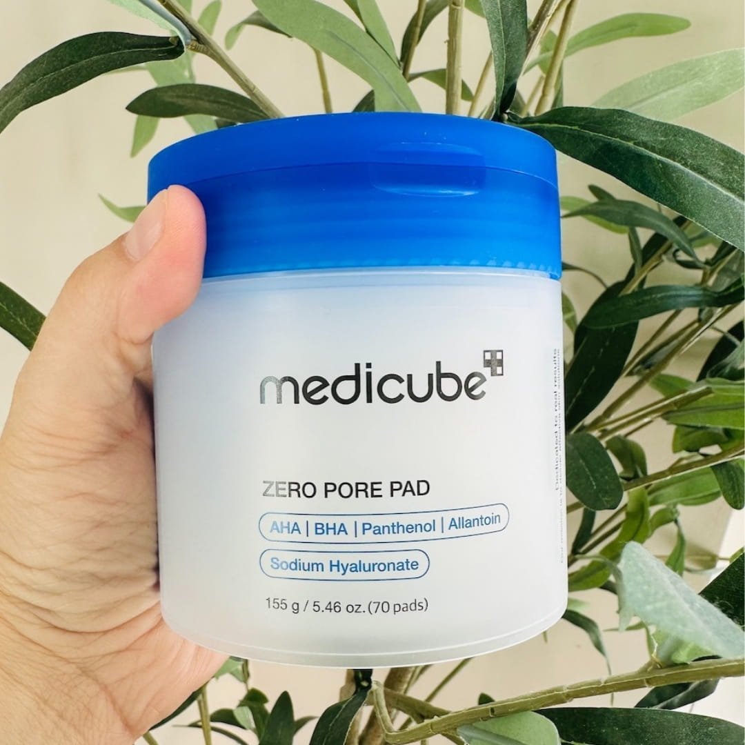 Medicube Zero Pore Pad toner pads in a white cylindrical container with a blue lid, featuring AHA, BHA, Panthenol, and Sodium Hyaluronate, held by a hand against a natural green leafy background.