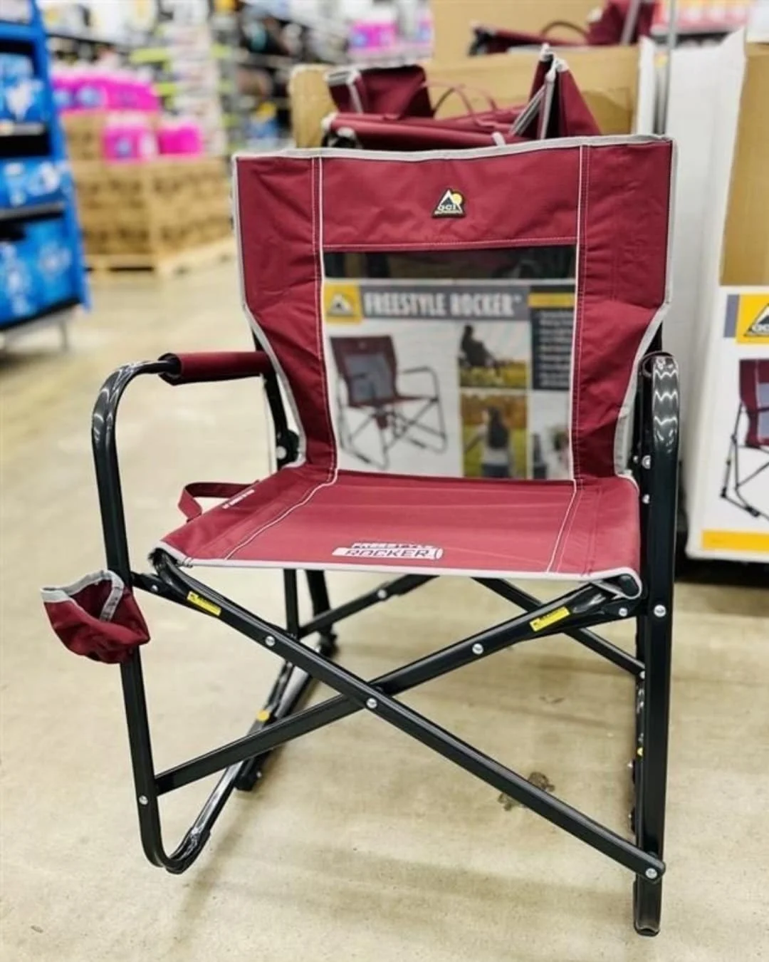 GCI Outdoor Freestyle Rocker folding chair in burgundy and grey with a mesh backrest, padded armrests, and a cup holder, displayed in a retail warehouse setting.