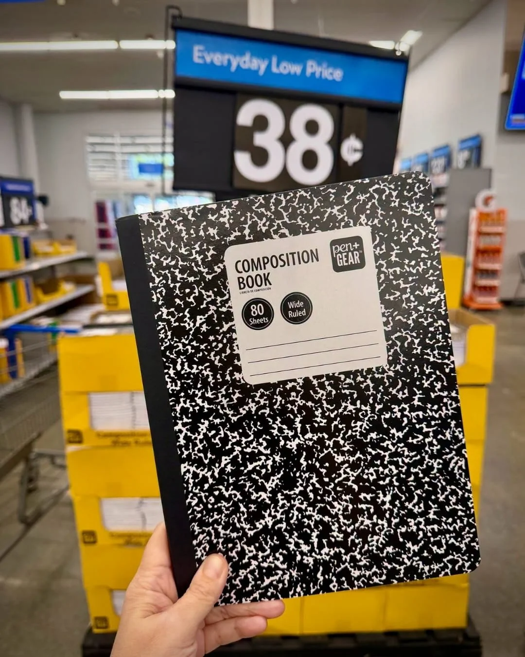 Hand holding a Pen+Gear wide-ruled composition book with a black marble cover in a retail store aisle
