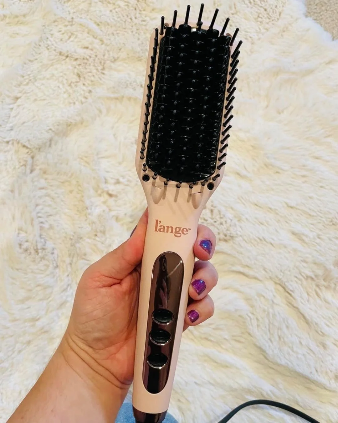 Blush pink L'ange thermal straightening brush with black bristles and a metallic control panel, held by a hand with purple glittery nails against a cream faux fur background.