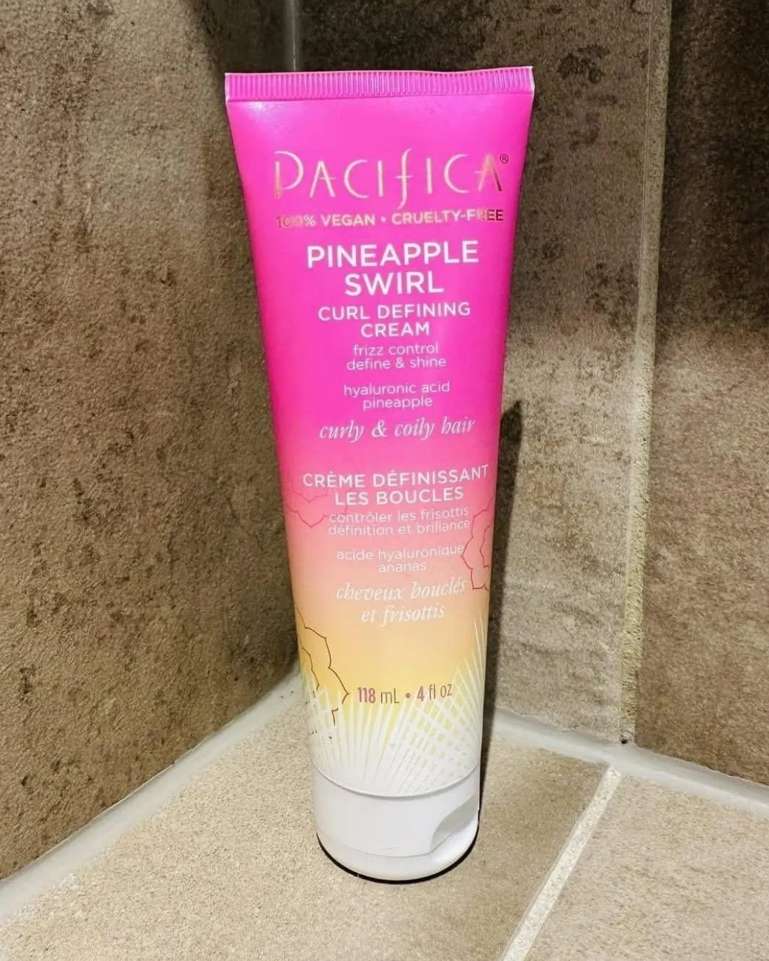 Tube of Pacifica Pineapple Swirl Curl Defining Cream with a vibrant pink-to-yellow gradient design, featuring text for frizz control and shine, placed on beige tiled bathroom surface.