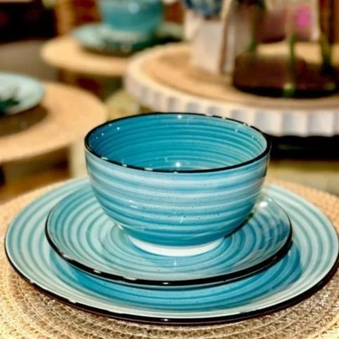 Close-up of a blue and white dish set with a swirling pattern, featuring a bowl, smaller plate, and larger plate stacked on a woven placemat, with blurred background elements of similar tableware on wooden stands.