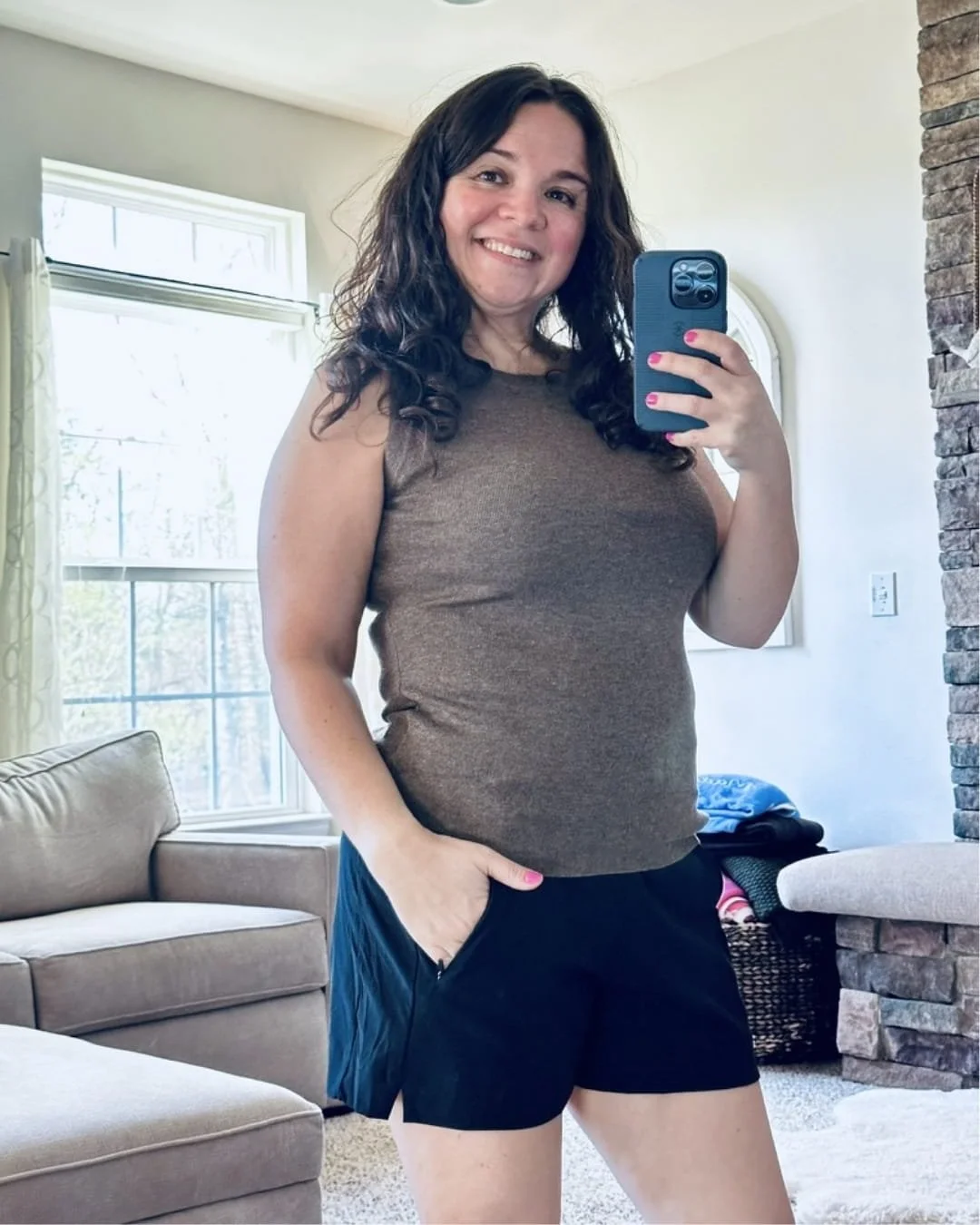 Nicole taking a mirror selfie in a brown ribbed Arach & Cloz tank top and black athletic shorts, standing in a well-lit living room with neutral-toned furniture and a stone accent wall.