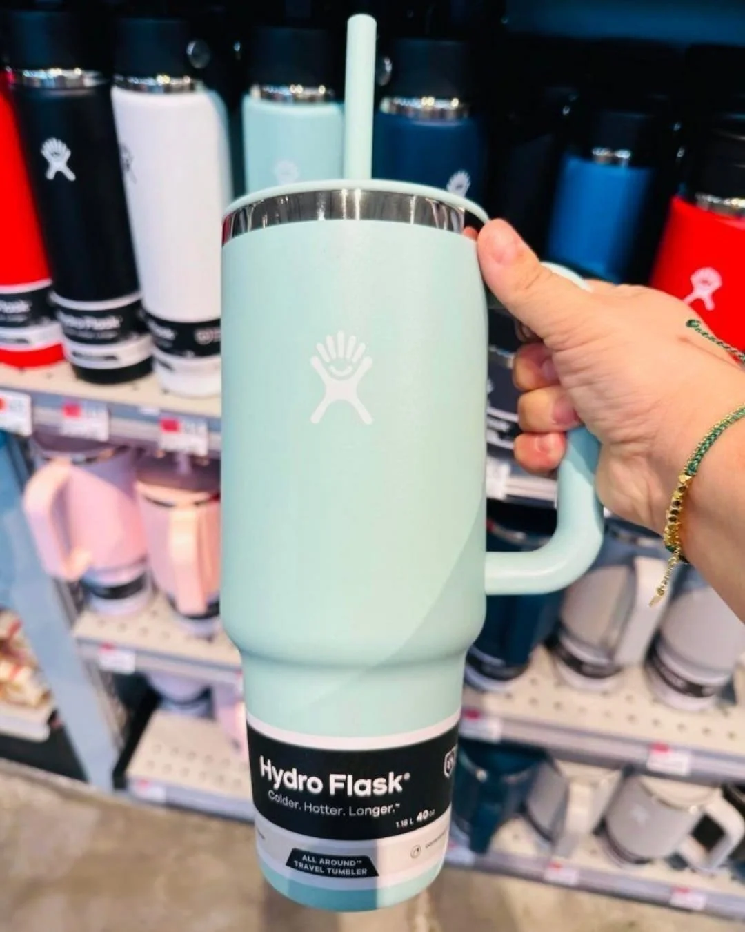 Mint green Hydro Flask 40 oz All Around Travel Tumbler with handle and straw, held by a hand in front of a retail display of Hydro Flask bottles in various colors.