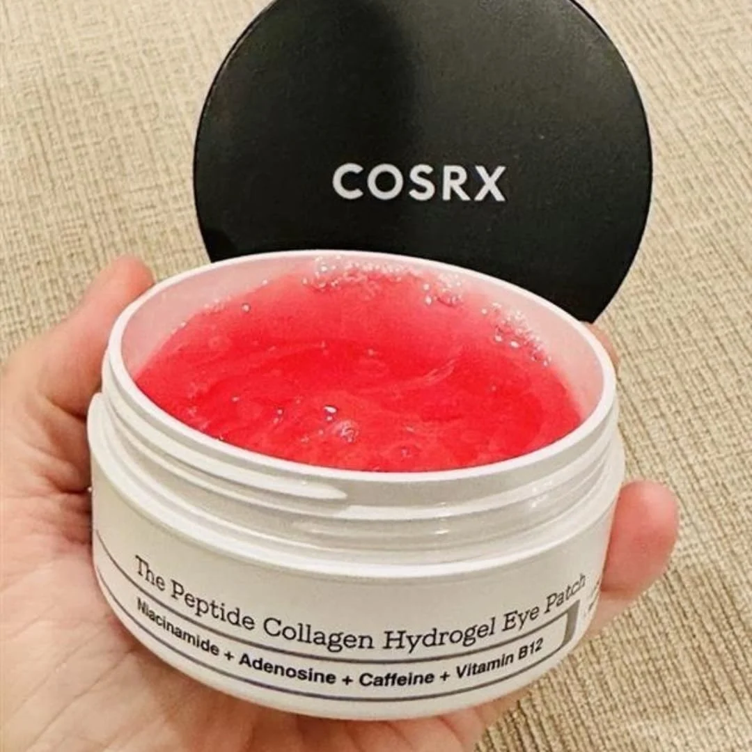 Close-up of COSRX Peptide Collagen Hydrogel Eye Patch jar held in a hand, showcasing red hydrogel patches with key ingredients like niacinamide, adenosine, caffeine, and vitamin B12.