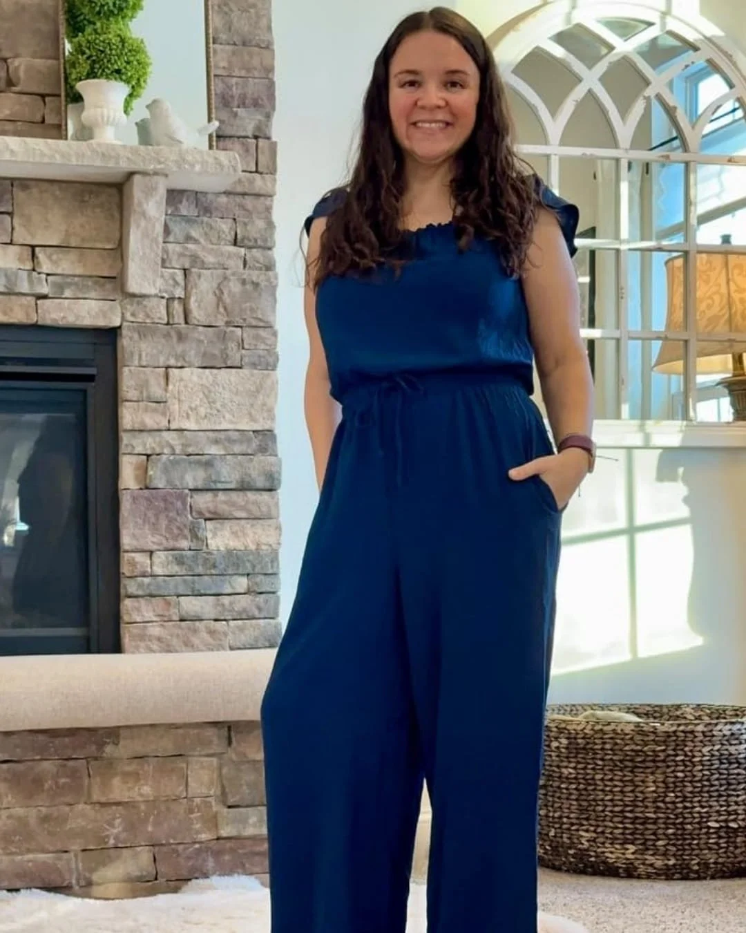 Nicole wearing a navy blue sleeveless jumpsuit with ruffled neckline and wide-leg trousers, standing indoors in a bright living room with a stone fireplace, arched window, and natural decor accents.