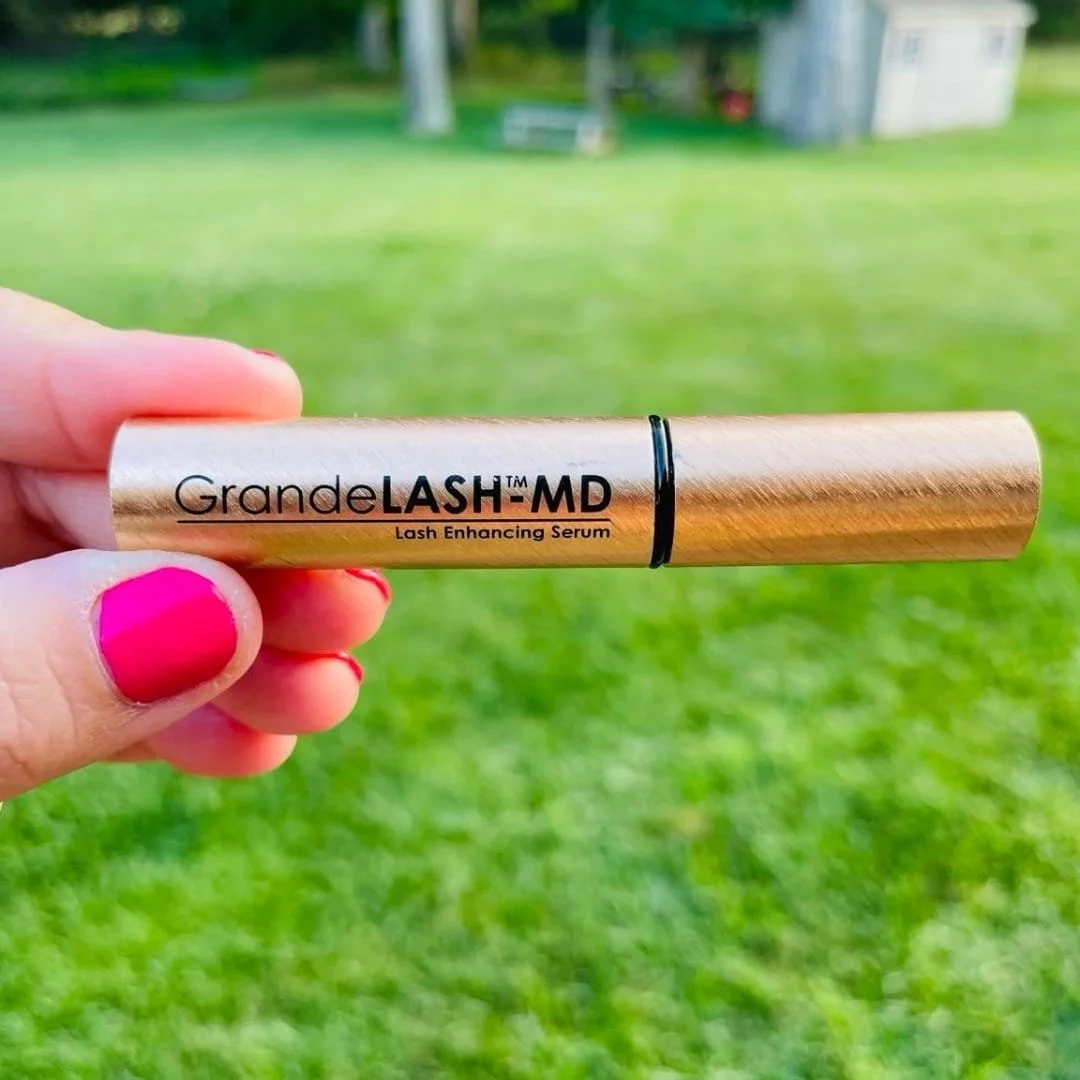 A metallic rose-gold tube of GrandeLASH-MD Lash Enhancing Serum held horizontally by a hand with bright pink nails, set against a blurred outdoor background with green grass and trees.