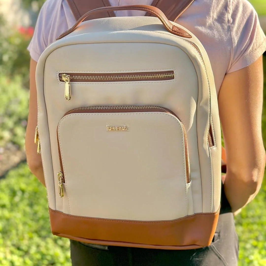 Telena leather laptop backpack in light cream with brown accents, gold zippers, and multiple compartments, worn outdoors against a green foliage background.