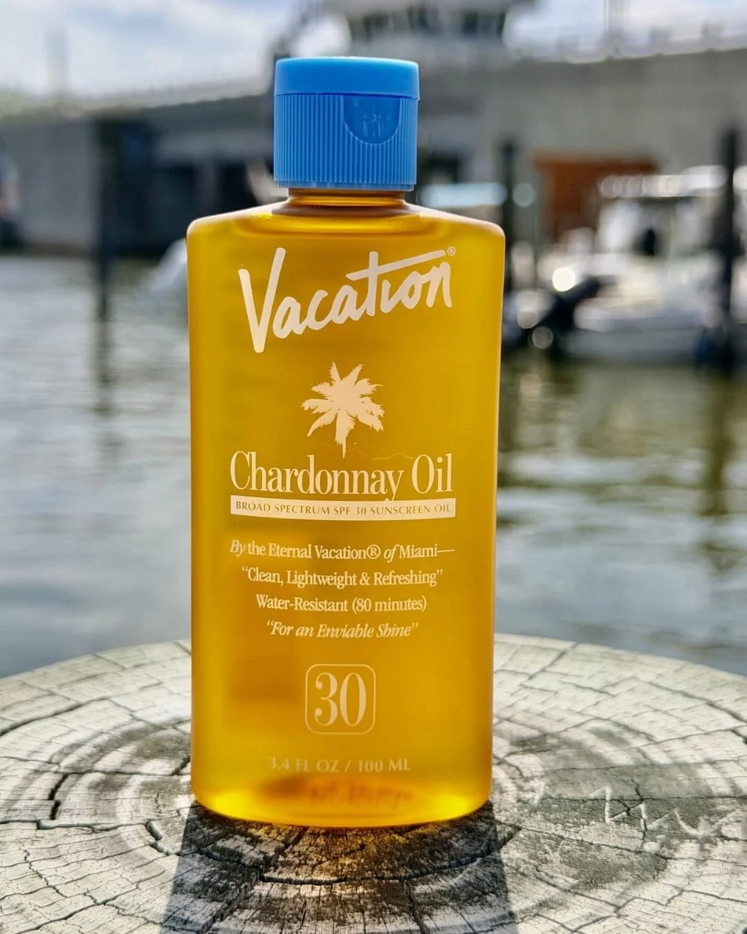 Bottle of Vacation Chardonnay Oil SPF 30 sunscreen oil displayed on a weathered wooden post by the water