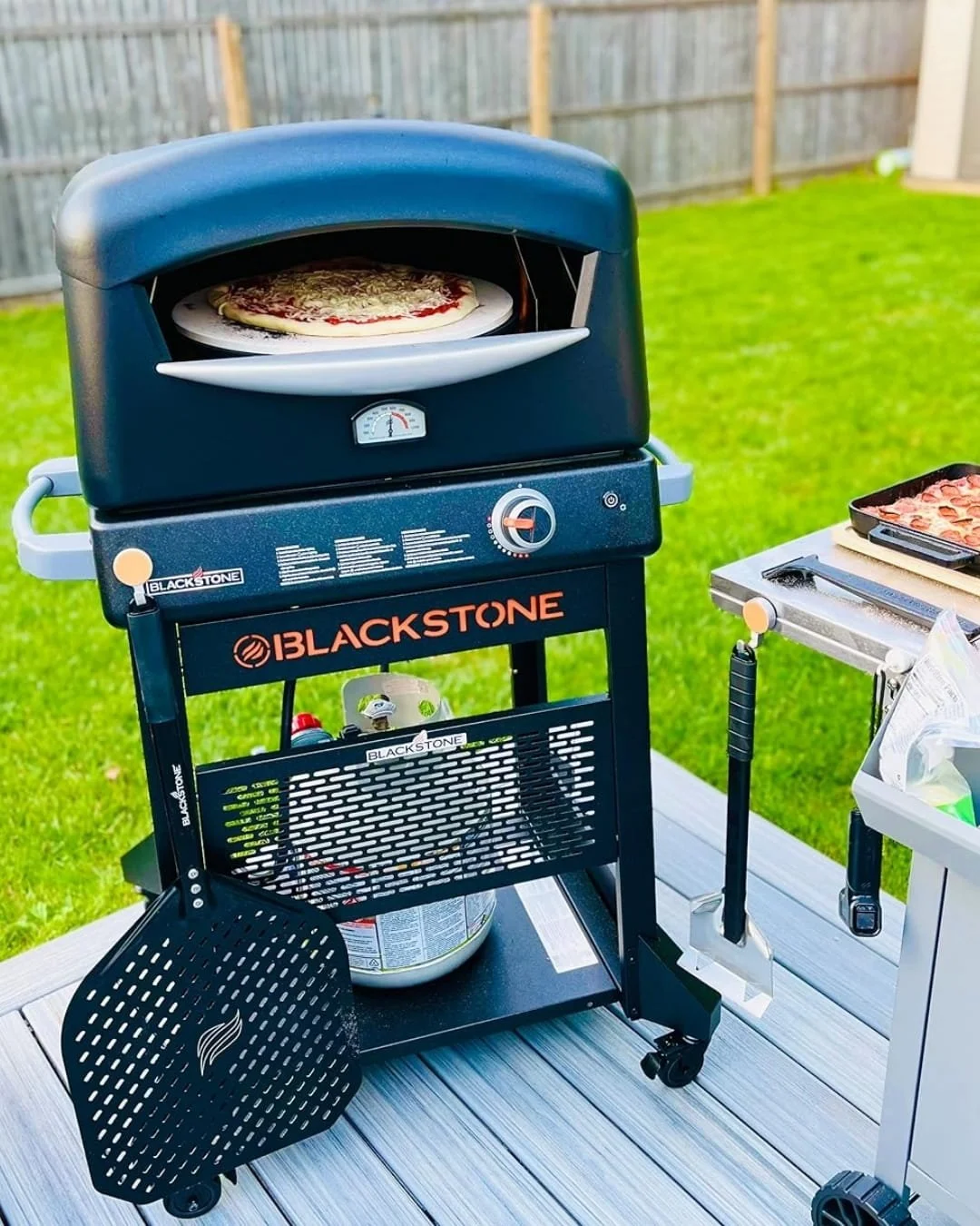 BakerStone outdoor pizza oven with a pizza cooking on a stone inside, set on a backyard deck with a prep table holding a pepperoni pizza and cooking tools, surrounded by a green lawn and wooden fence.