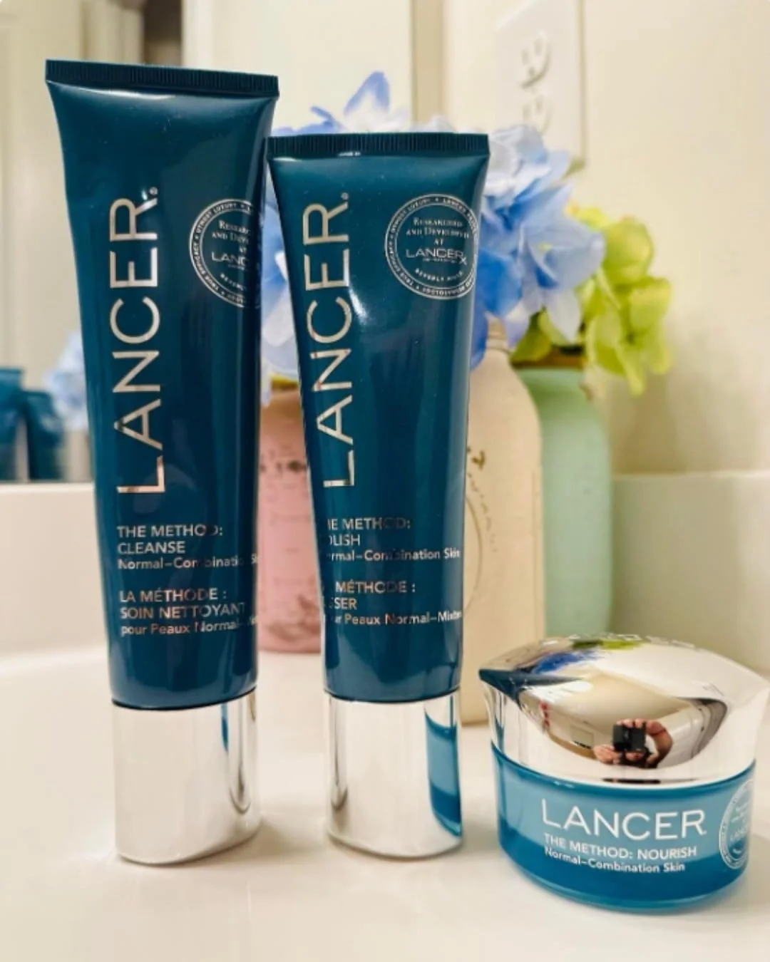 Lancer skincare kit with Cleanse, Polish, and Nourish products arranged on a white surface with soft floral accents in the background