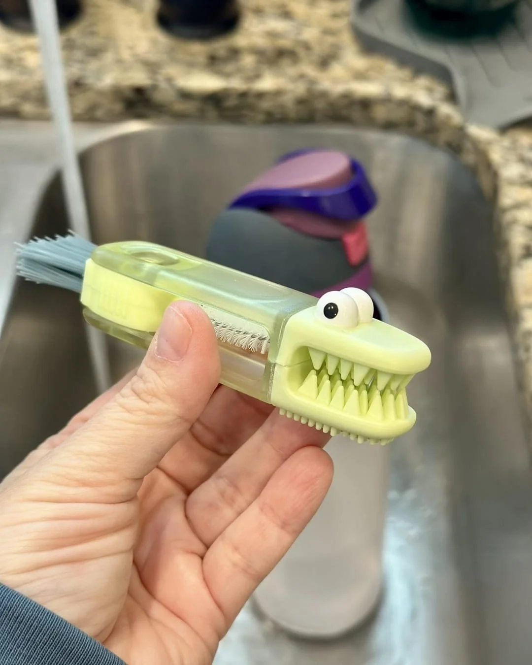 Hand holding a green alligator-shaped 3-in-1 cleaning tool with a silicone scrubber head, spiral brush, and tail bristles, designed for cleaning cup lids and gaps, shown over a stainless steel kitchen sink.