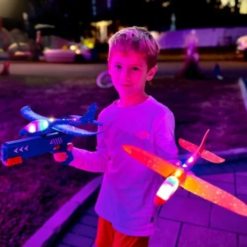 Young boy outdoors at night holding two light-up toy airplanes, one blue on a launcher and one orange, illuminated by vibrant purple lighting with a blurred dark background.