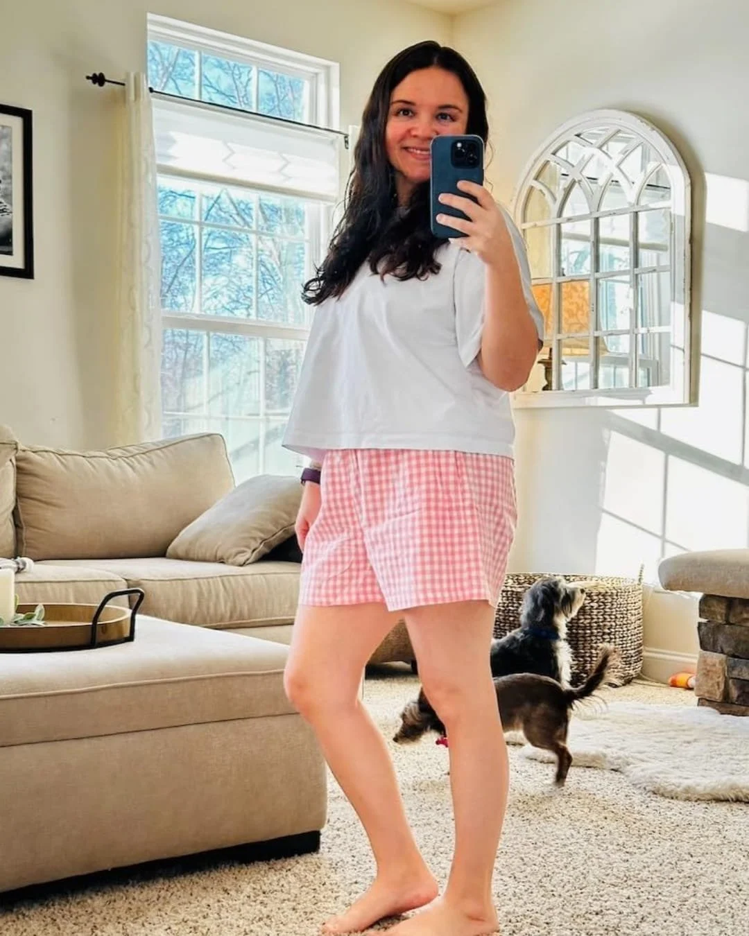 Nicole in white t-shirt and pink gingham boxer shorts taking a mirror selfie in a bright living room with a beige sectional sofa, two small dogs, and natural light streaming through large windows.