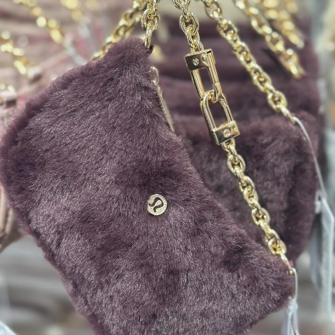 Lululemon faux-fur shoulder bag in deep purple with a gold chain strap and circular gold logo, displayed against a blurred retail background.