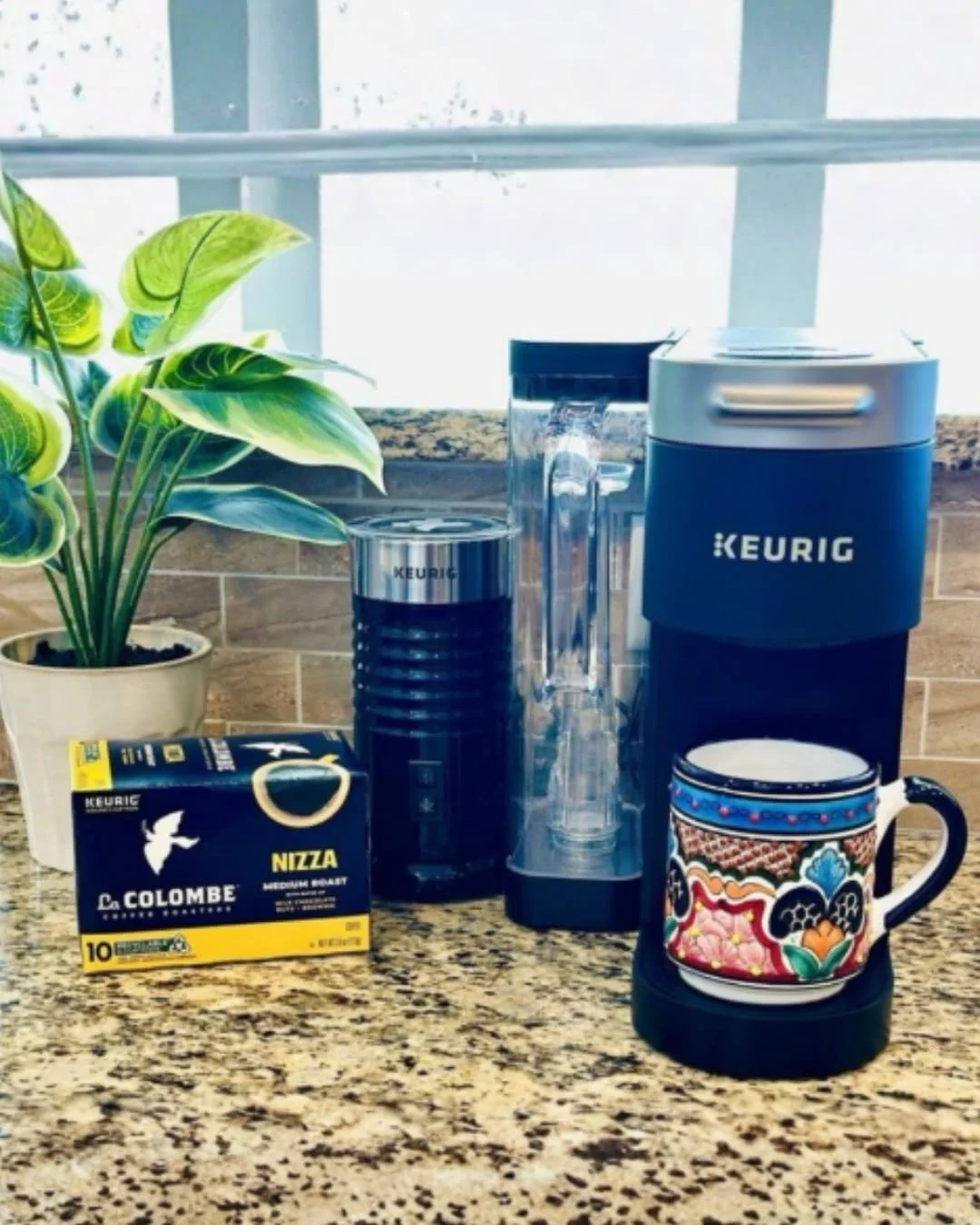 Keurig coffee station featuring a slim navy coffee maker, black milk frother, and a box of La Colombe Nizza medium roast K-Cup pods, set on a granite countertop with a potted plant and a decorative ceramic mug in a modern kitchen.