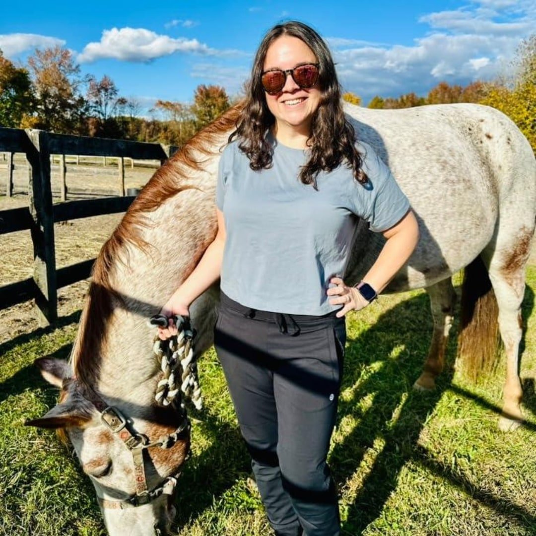Nicole, CEO of Nicole Fabulous Finds, smiling outdoors with a spotted horse in a sunny autumn pasture, featuring a wooden fence and fall-colored trees in the background.