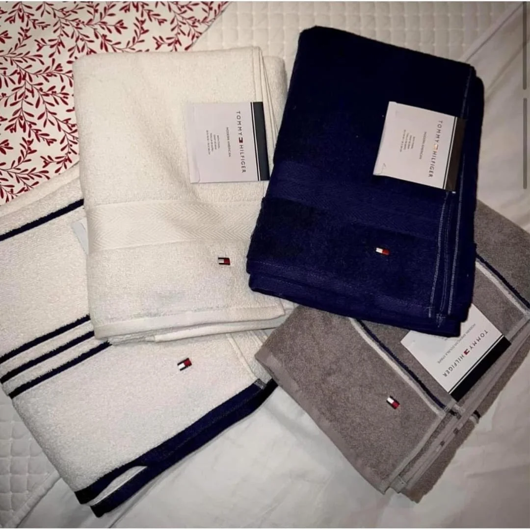 Tommy Hilfiger bath towel collection featuring folded towels in white, navy blue, and gray with embroidered logo and stripe patterns, displayed on white bedding.