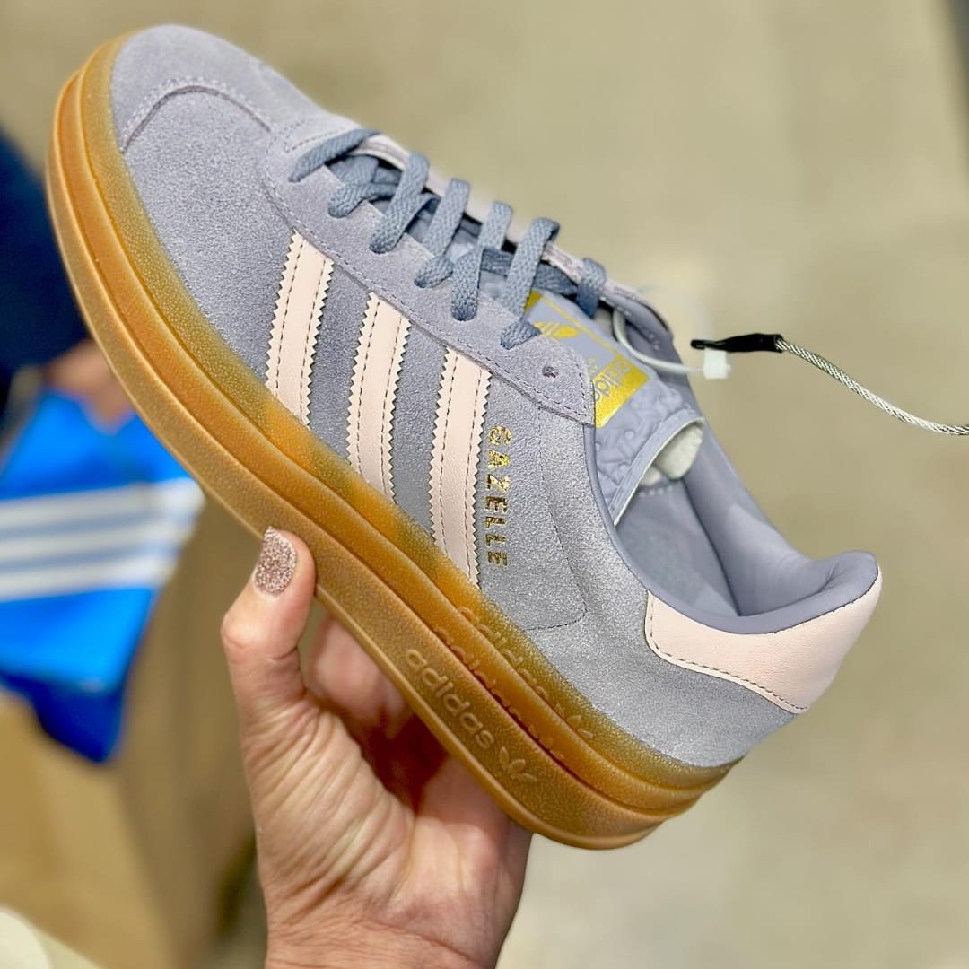 Adidas Gazelle sneaker with light gray suede upper, blush pink stripes and heel tab, gold GAZELLE lettering, and gum-colored platform sole, held by a hand with glittery nail polish against a blurred indoor background.