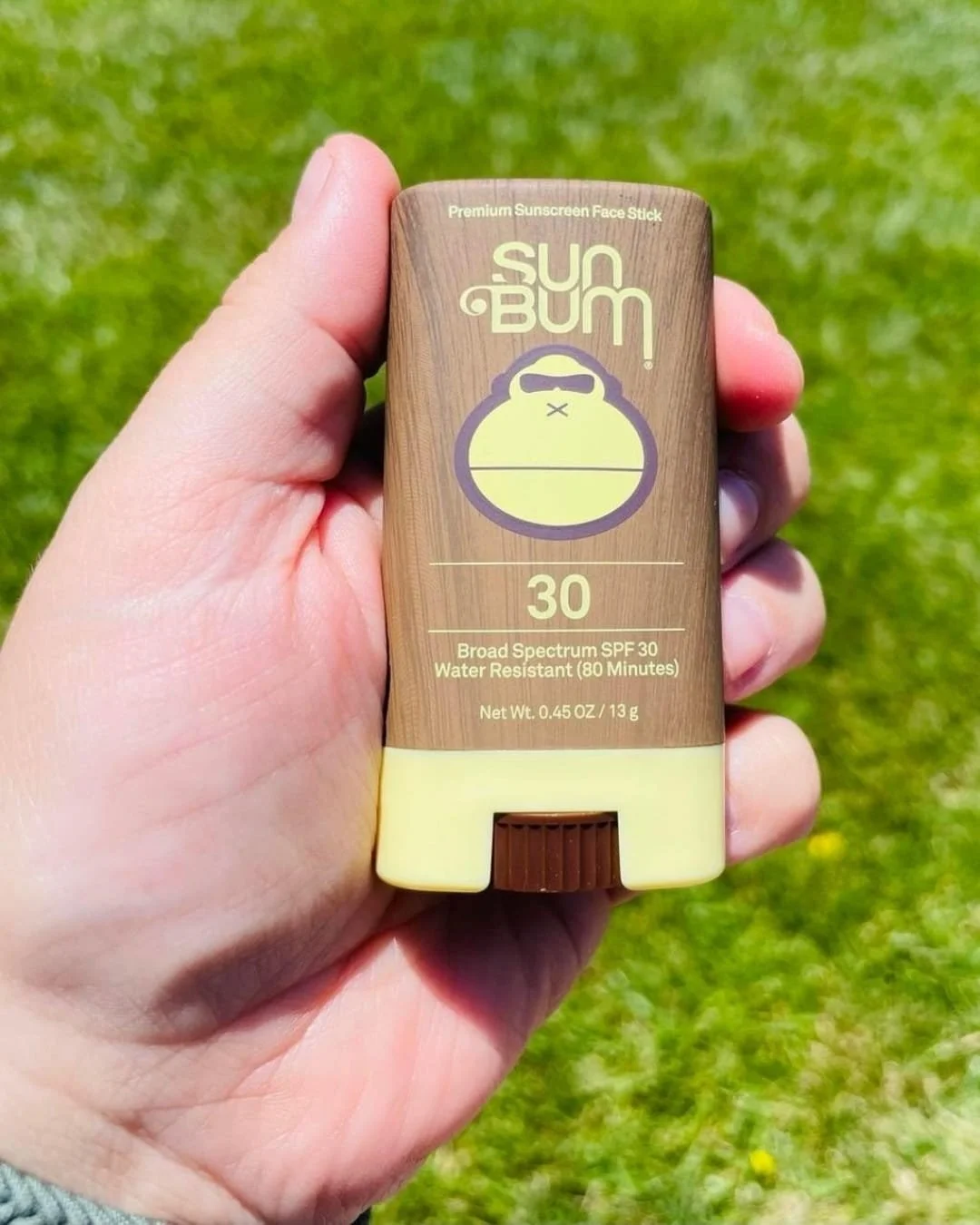 Sun Bum Face Sunscreen Stick SPF 30 held in hand outdoors against green grass background