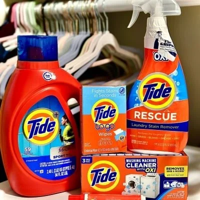 Display of Tide laundry products including a red bottle of liquid detergent, a box of Tide to go wipes, a spray bottle of Tide Rescue stain remover, and a box of Tide Washing Machine Cleaner, set in front of a clothes rack with hangers.
