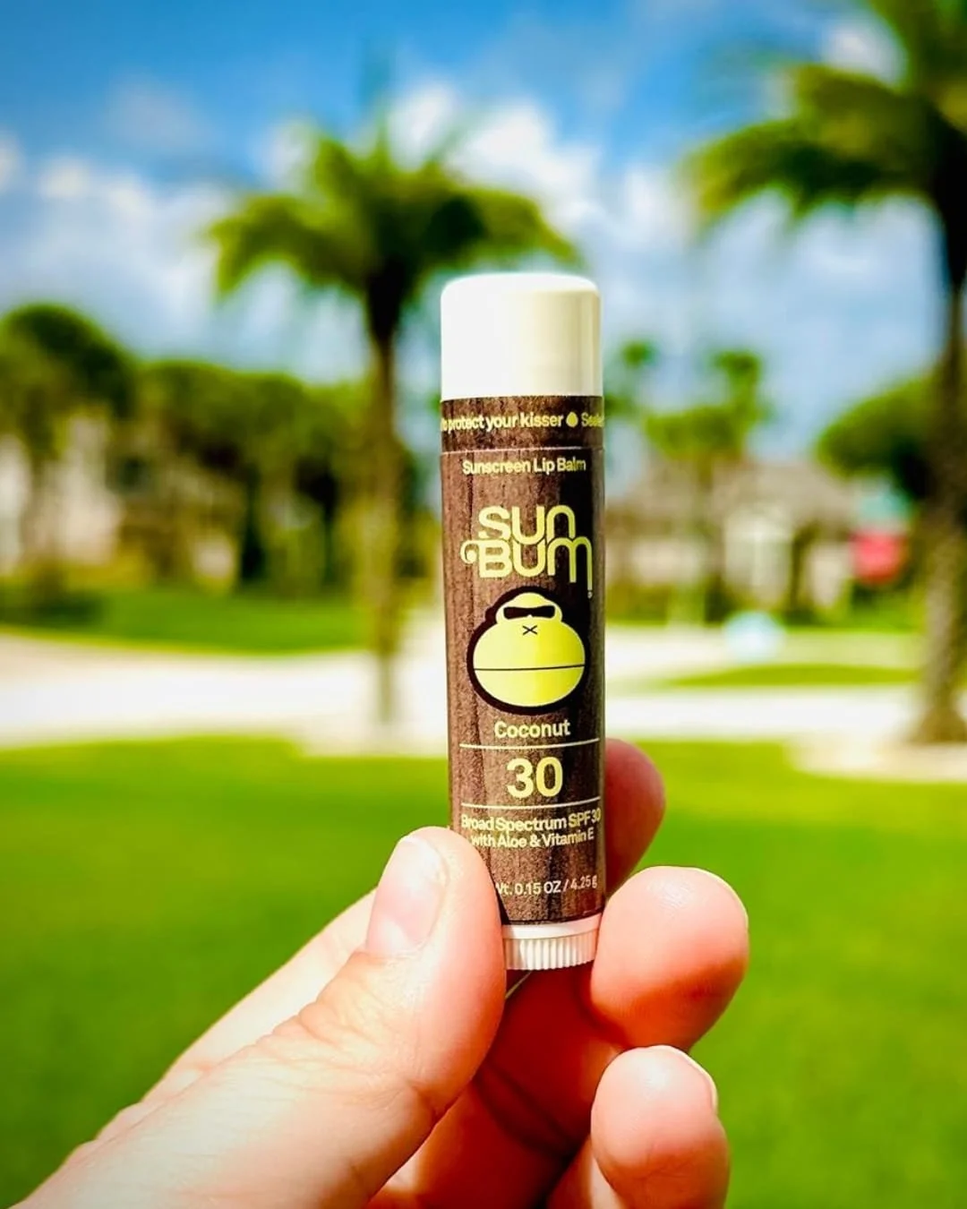 Sun Bum Coconut Sunscreen Lip Balm (SPF 30) held vertically in a hand against a sunny tropical background with blurred palm trees, green grass, and a bright blue sky.