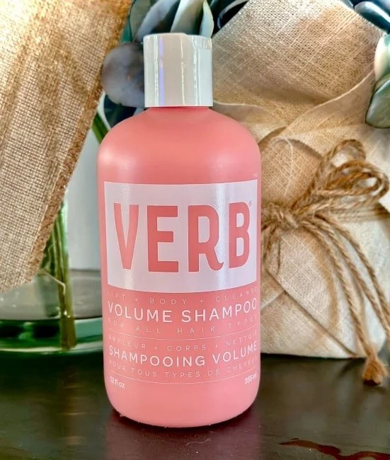 VERB Volume Shampoo in a pink bottle with white label, featuring lift + body + cleanse benefits, styled with green foliage and burlap decor on a wooden surface, perfect for all hair types.