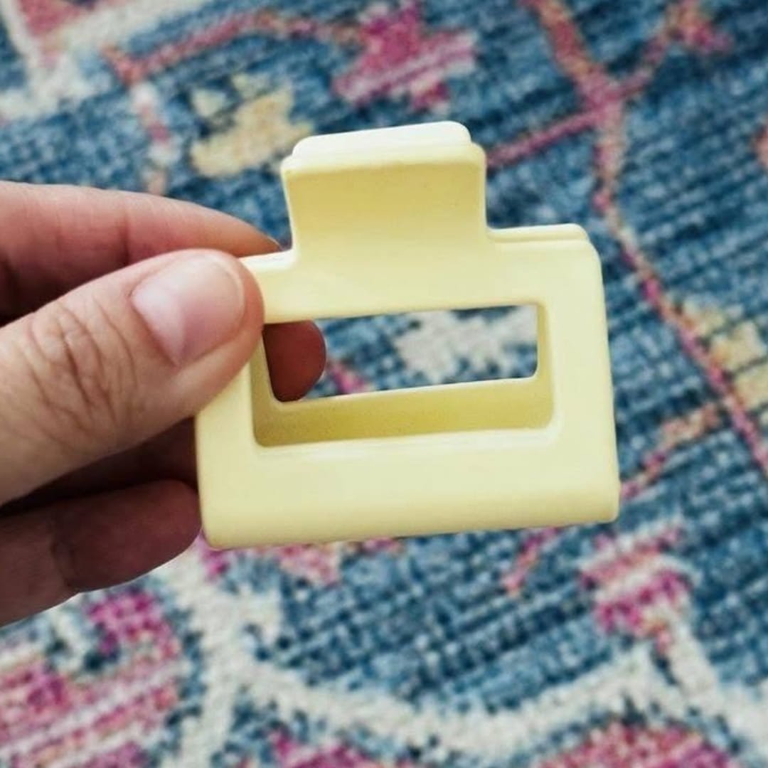 Hand holding a pale yellow geometric women’s hair claw clip with a rectangular cutout, shown against a textured blue and pink patterned rug background