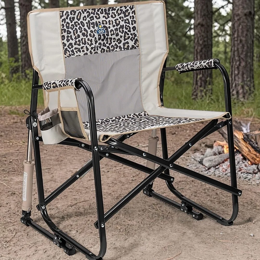GCI Rocker XL camp chair with leopard-print accents, beige canvas, and a mesh backrest, featuring a rocking mechanism and side storage pockets, set at a campsite with a fire pit and pine trees in the background.