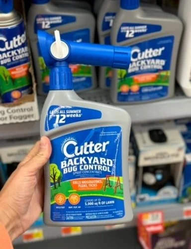 Hand holding a Cutter Backyard Bug Control Spray Concentrate bottle with a blue sprayer, featuring 12 weeks and kills mosquitoes, fleas, ticks on the label, with similar products visible on a store shelf in the background.