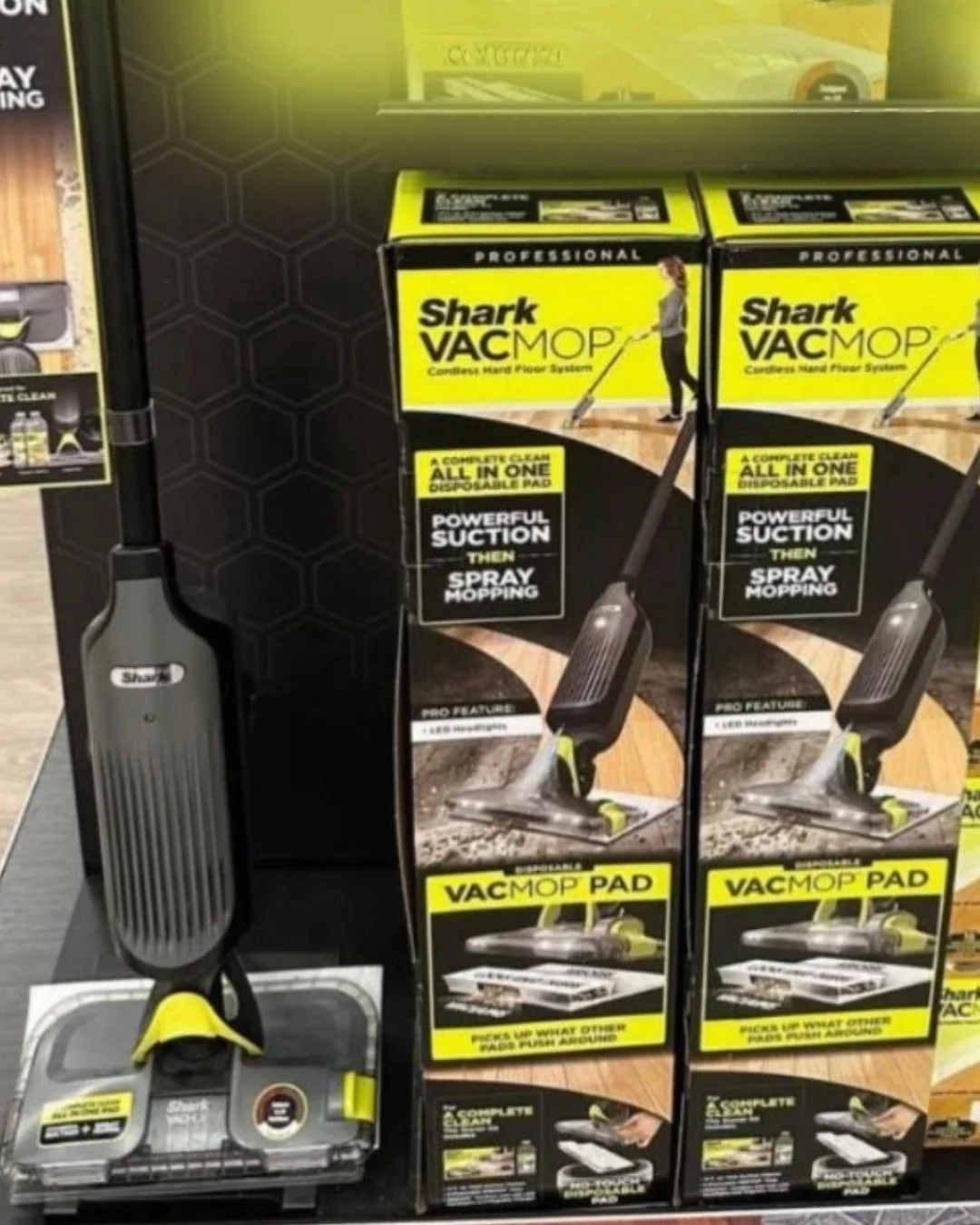 Retail display of Shark VACMOP Cordless Hard Floor System, featuring a demo unit on a black stand and two product boxes highlighting features like disposable pads, powerful suction, and spray mopping.