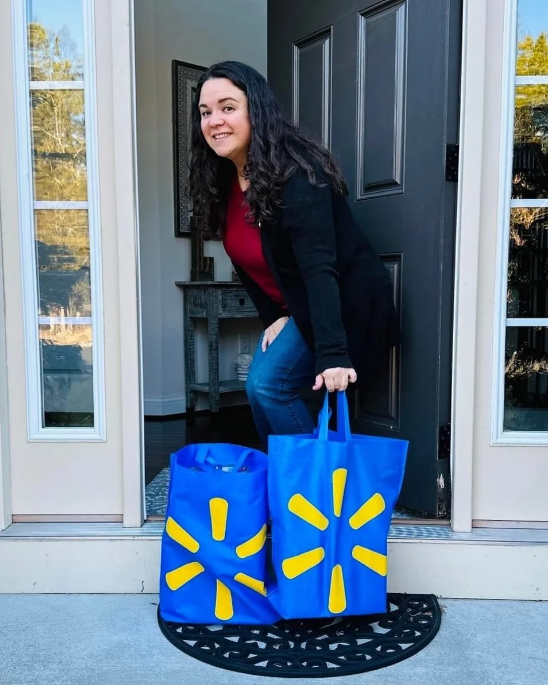 Nicole smiling at her open front door, holding two blue Walmart reusable shopping bags with the yellow spark logo, showcasing a convenient home delivery or shopping experience.