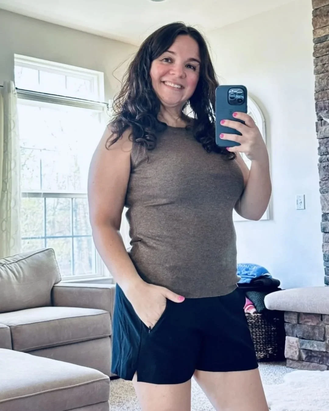 Nicole taking a mirror selfie in a brown ribbed Arach & Cloz tank top and black athletic shorts, standing in a well-lit living room with neutral-toned furniture and a stone accent wall.