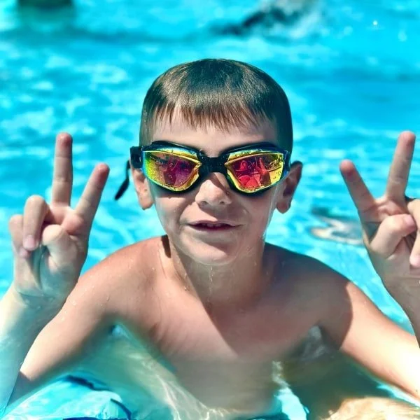 "Smiling boy in pool wearing goggles, making peace signs.