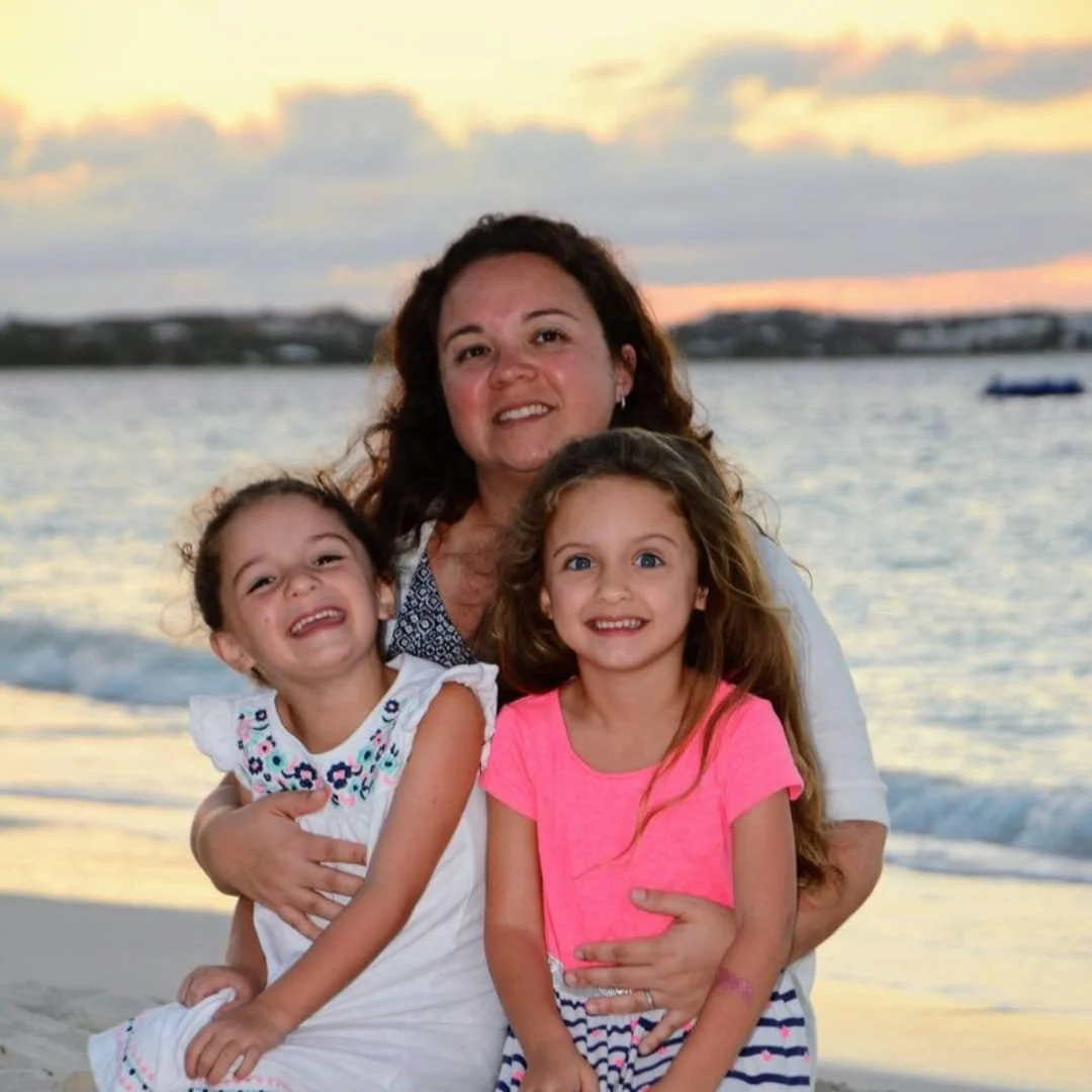 Nicole with her twin daughters smiling on a sandy beach during sunset, with a vibrant sky and calm sea in the background.